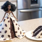 A doll with a dark brown complexion, African-inspired hairstyle, and a polka dot gown integrated into a matching doll-shaped cake. A slice of the cake is on a plate beside it, set in a bright kitchen with stainless steel appliances.