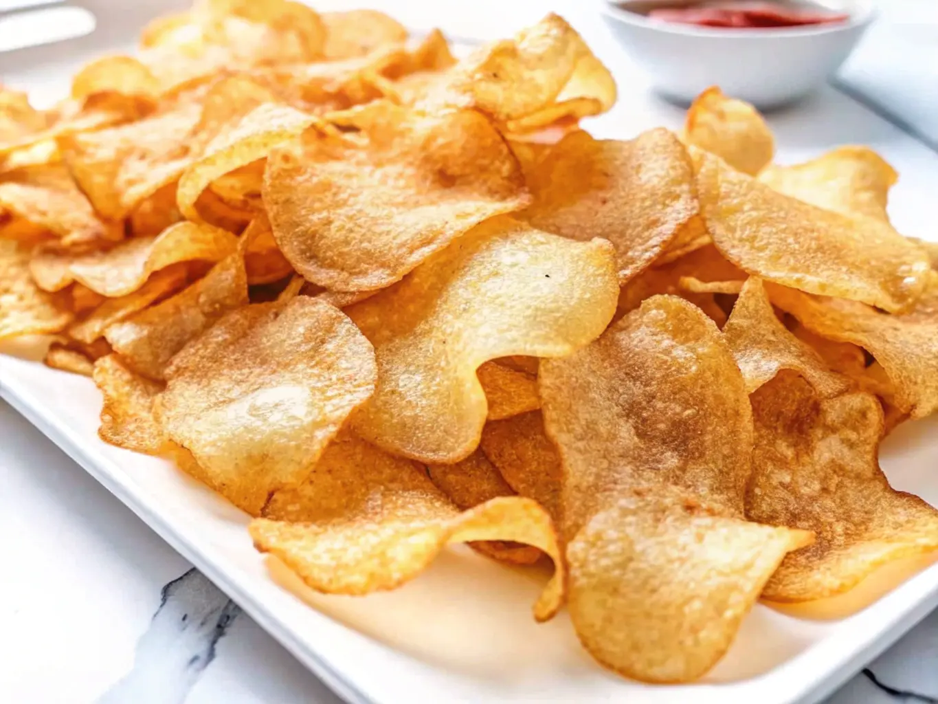 A plate of homemade salt and vinegar chips with a golden, crispy texture, served with a small bowl of ketchup in the background.