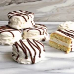 Bite-sized zebra cakes with creamy white filling and chocolate drizzle, displayed on a marble-tiled surface, with one partially sliced to reveal its layered interior.